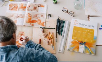Einem Herr sieht man über die Schulter beim malen mit Kreidefarben inspiriert von einem orangen Tontopf Kreide-Bild. Daneben sind noch einige Malutensilien zu sehen