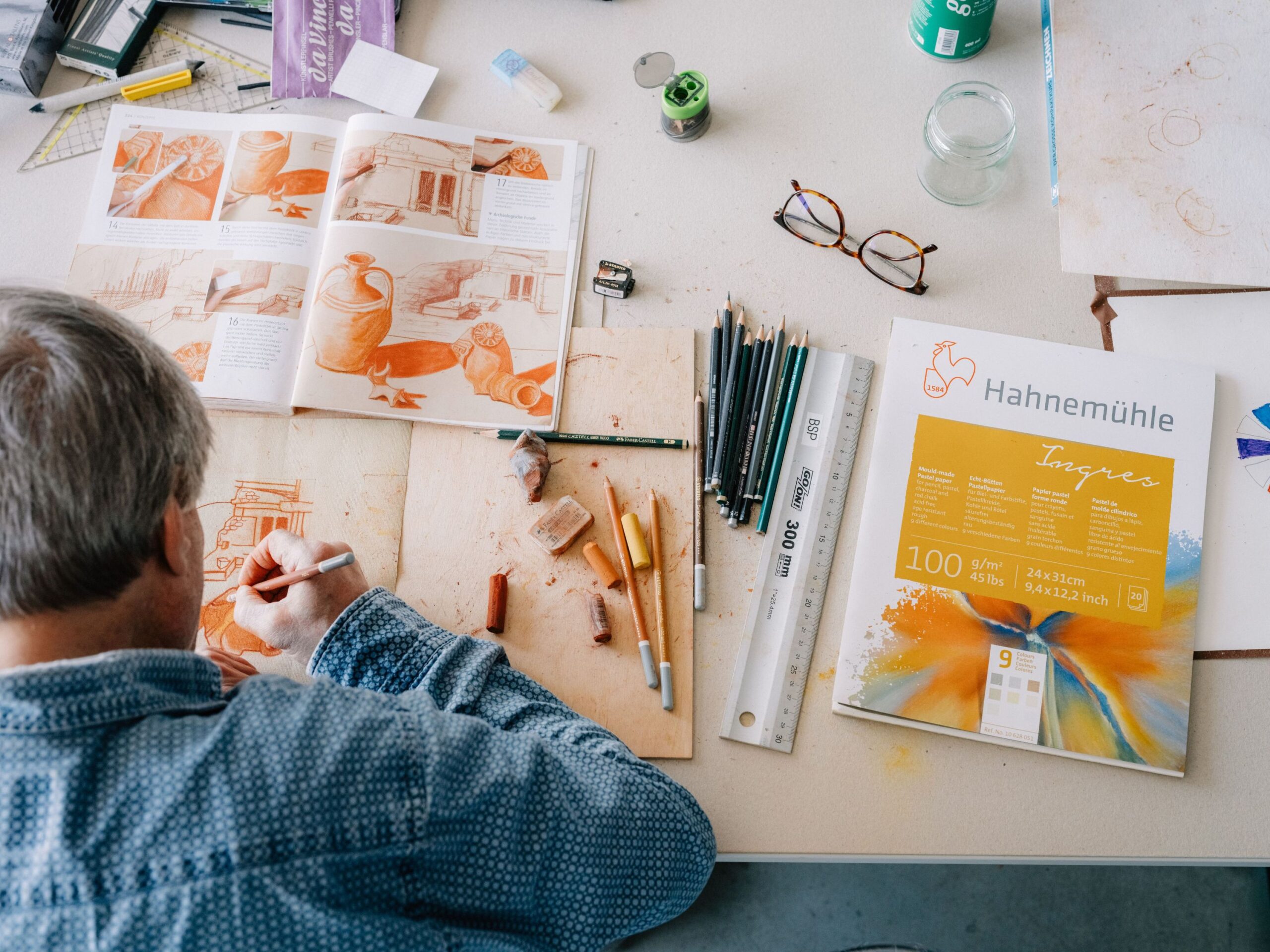 Einem Herr sieht man über die Schulter beim malen mit Kreidefarben inspiriert von einem orangen Tontopf Kreide-Bild. Daneben sind noch einige Malutensilien zu sehen